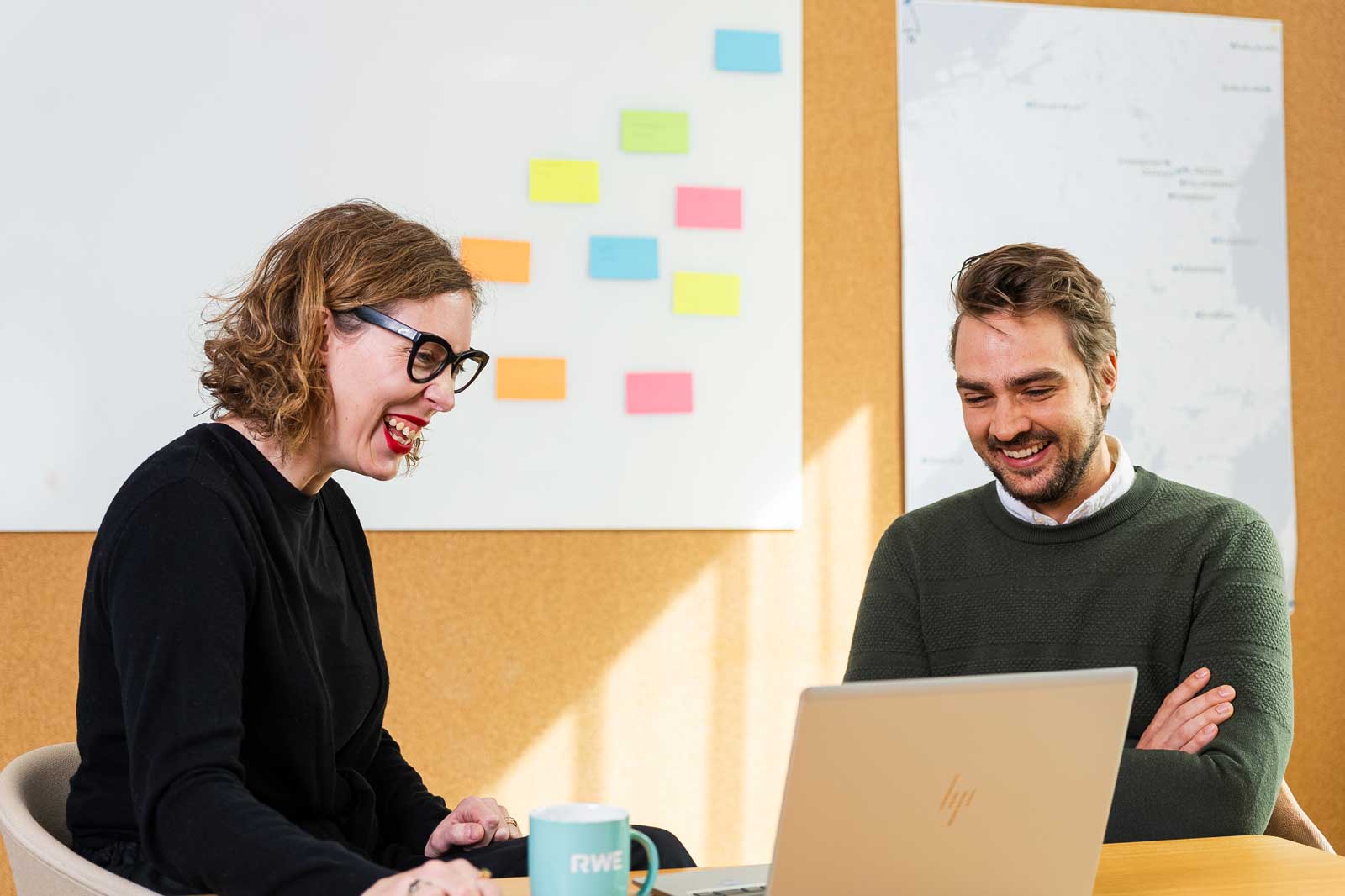 Two individuals engaged in discussion at a desk with a laptop and colourful sticky notes on the wall.