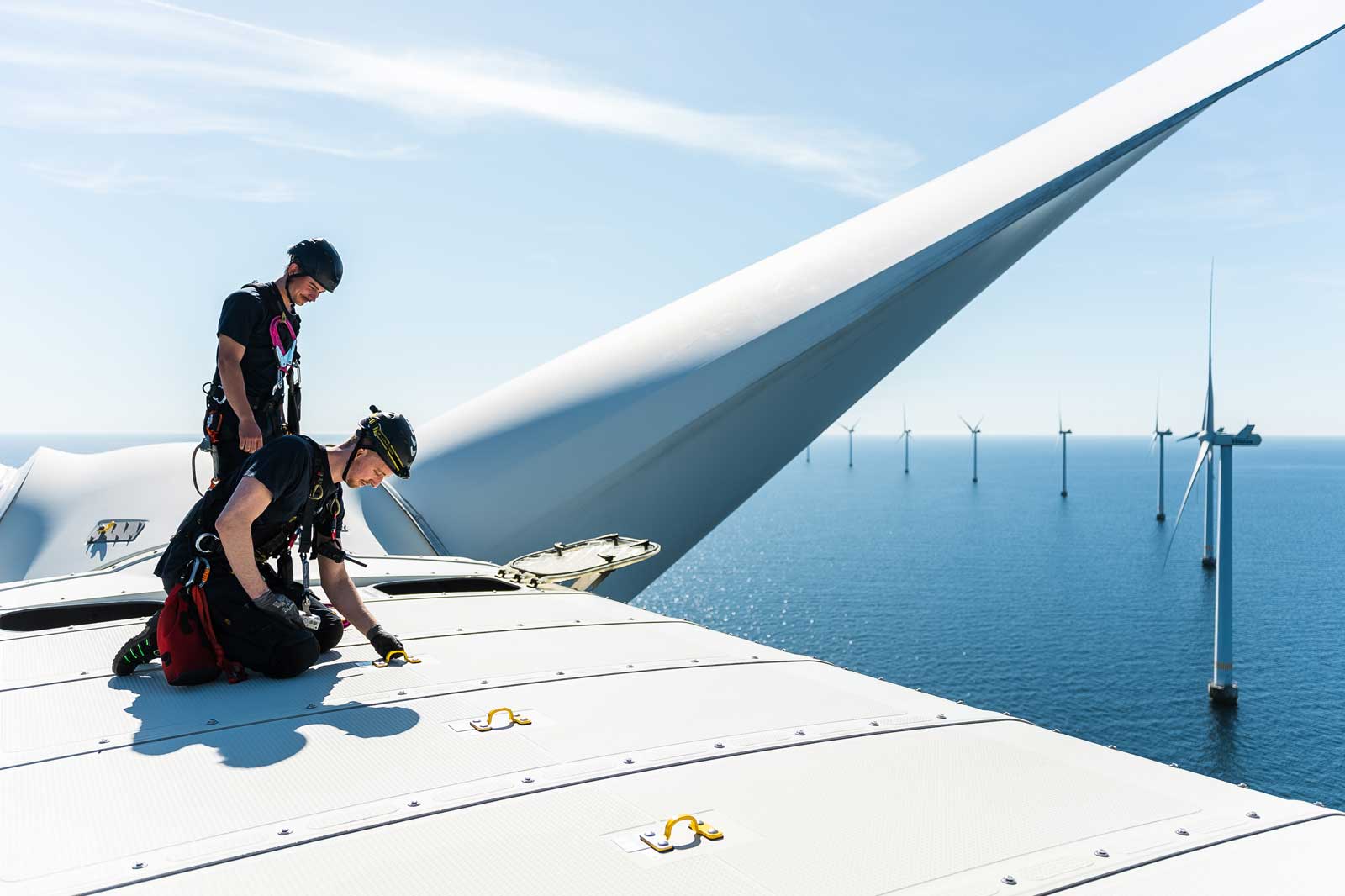 Two workers in safety gear on top of a wind turbine, overlooking offshore wind farms under a clear blue sky.