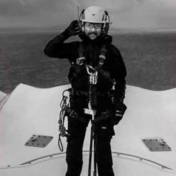 A person in a black harness and helmet stands on a boat deck, saluting against a backdrop of the sea and cloudy sky.