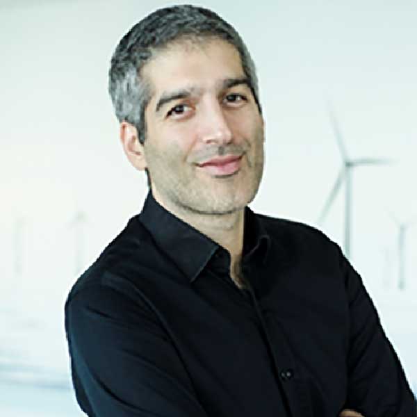 A person stands with crossed arms, wearing a black shirt, against a background of wind turbines.