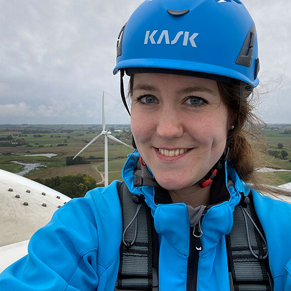 A person in a blue jacket and helmet stands on top of a wind turbine, overlooking a green landscape with fields and another turbine.