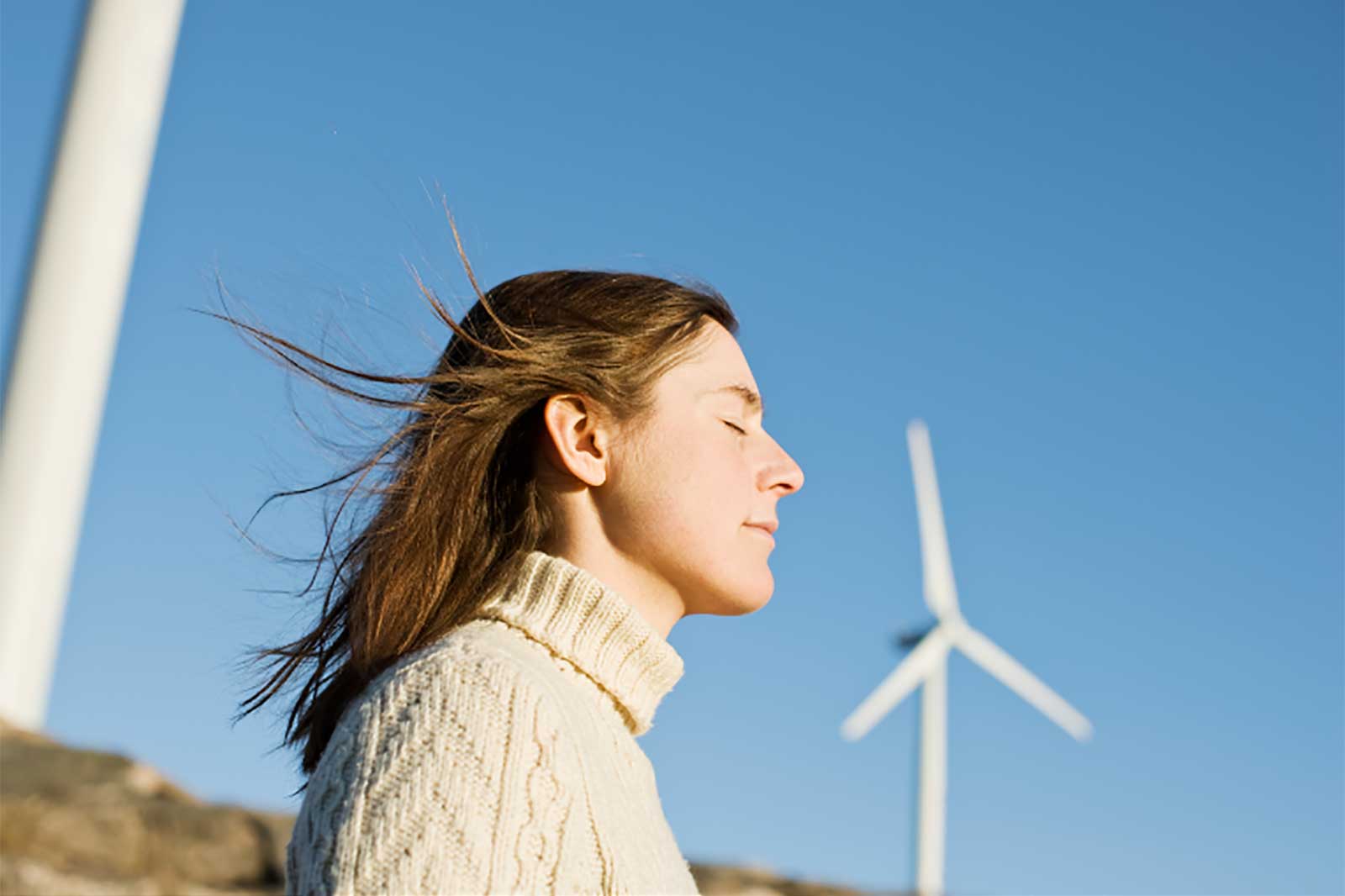 A person with flowing hair stands outdoors against a clear blue sky, with wind turbines visible in the background.