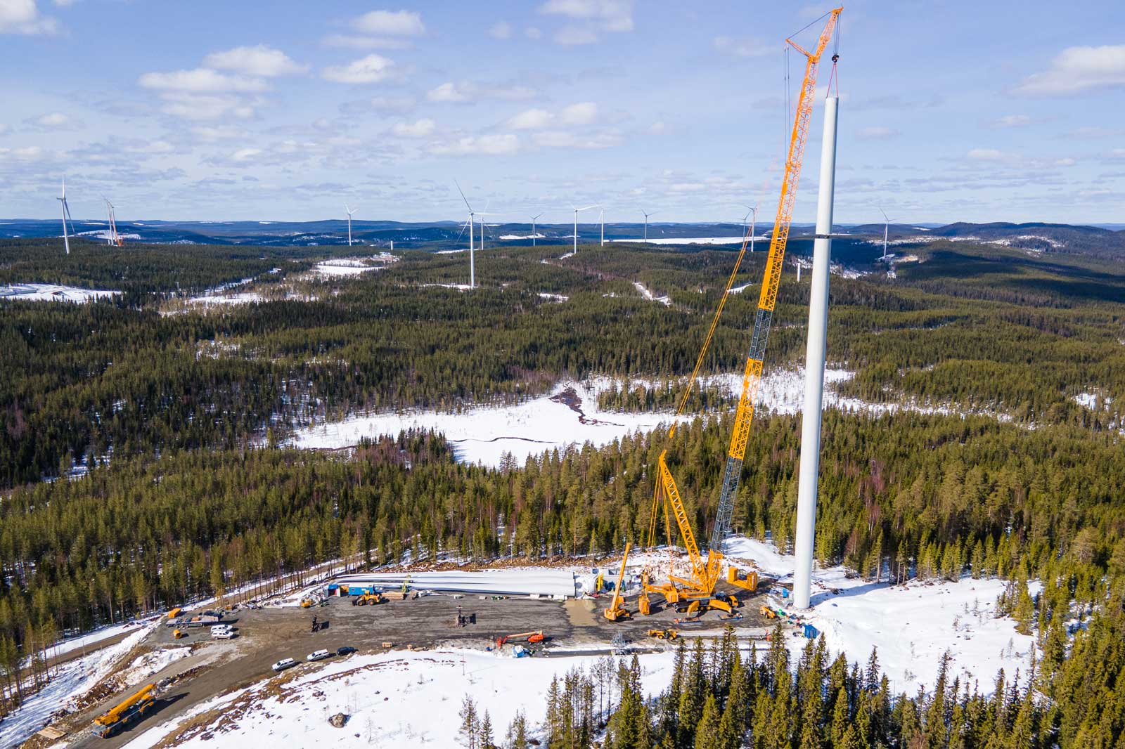 An aerial view of a wind turbine under construction in a snowy forest landscape, with machinery and equipment visible.