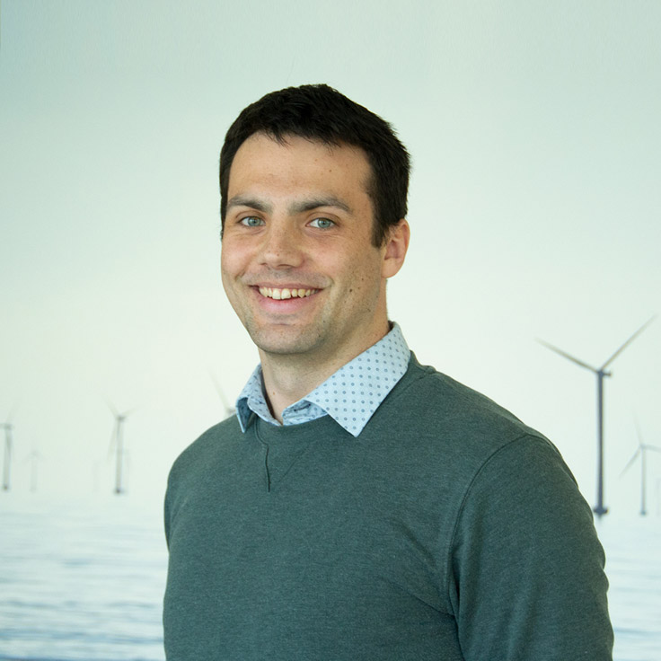 En man står leende framför en ljus bakgrund med vindkraftverk.