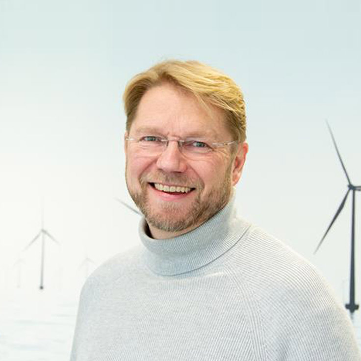 En person i en grå polotröja står mot en ljus bakgrund med vindkraftverk.