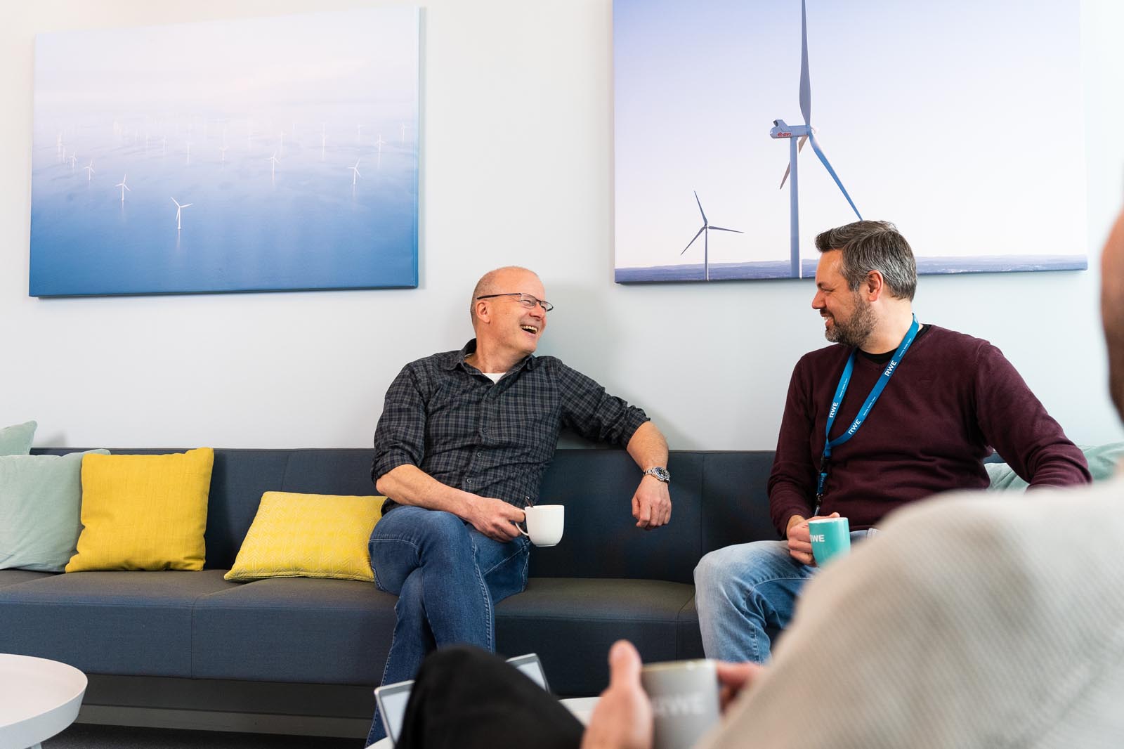 Två män diskuterar på en soffa, omgivna av dekorativa kuddar, med konstverk av vindkraftverk i bakgrunden.