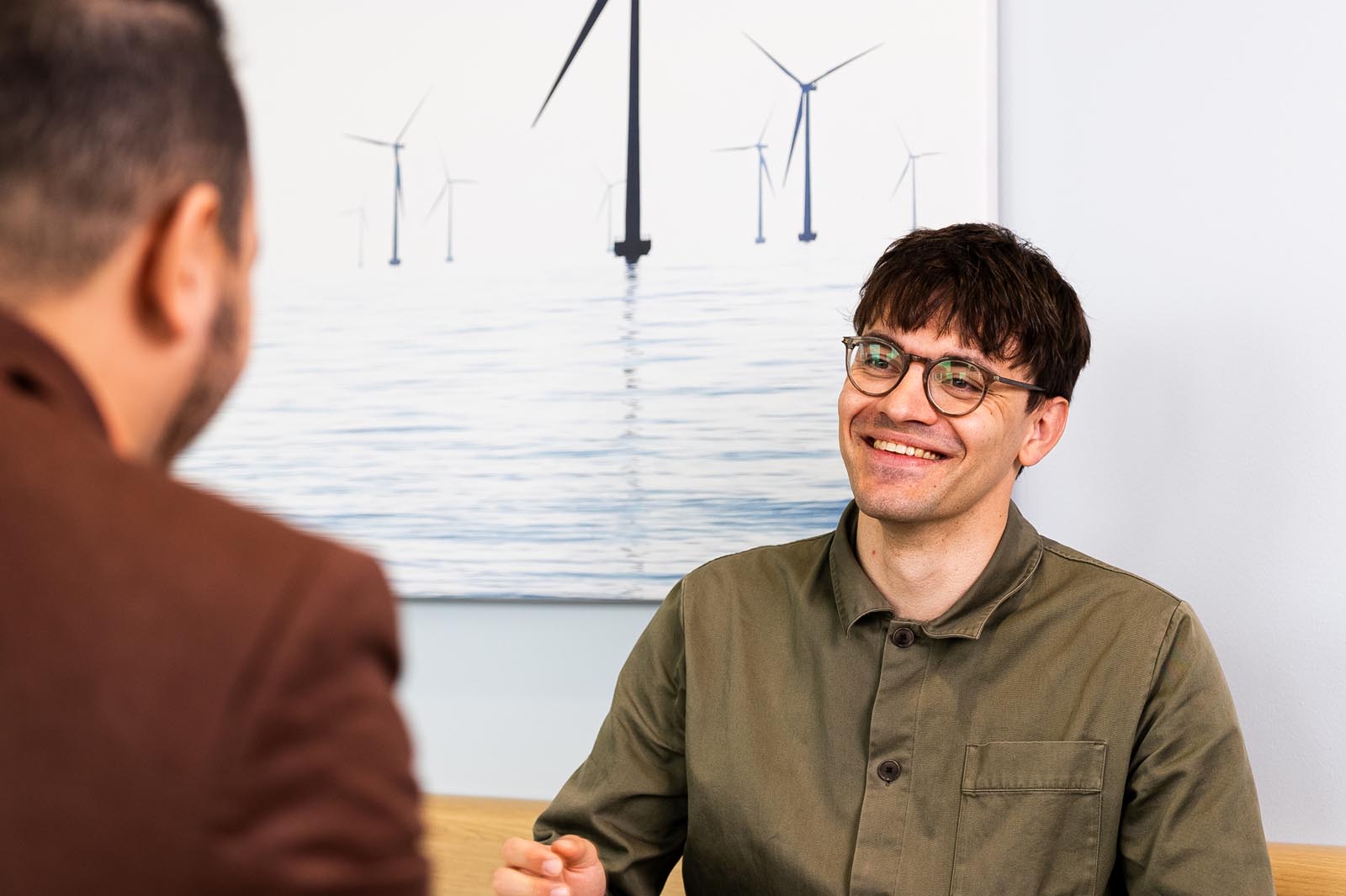 Ett samtal mellan två personer i en kontorsmiljö med en bakgrund av vindkraftverk och lugnt vatten.