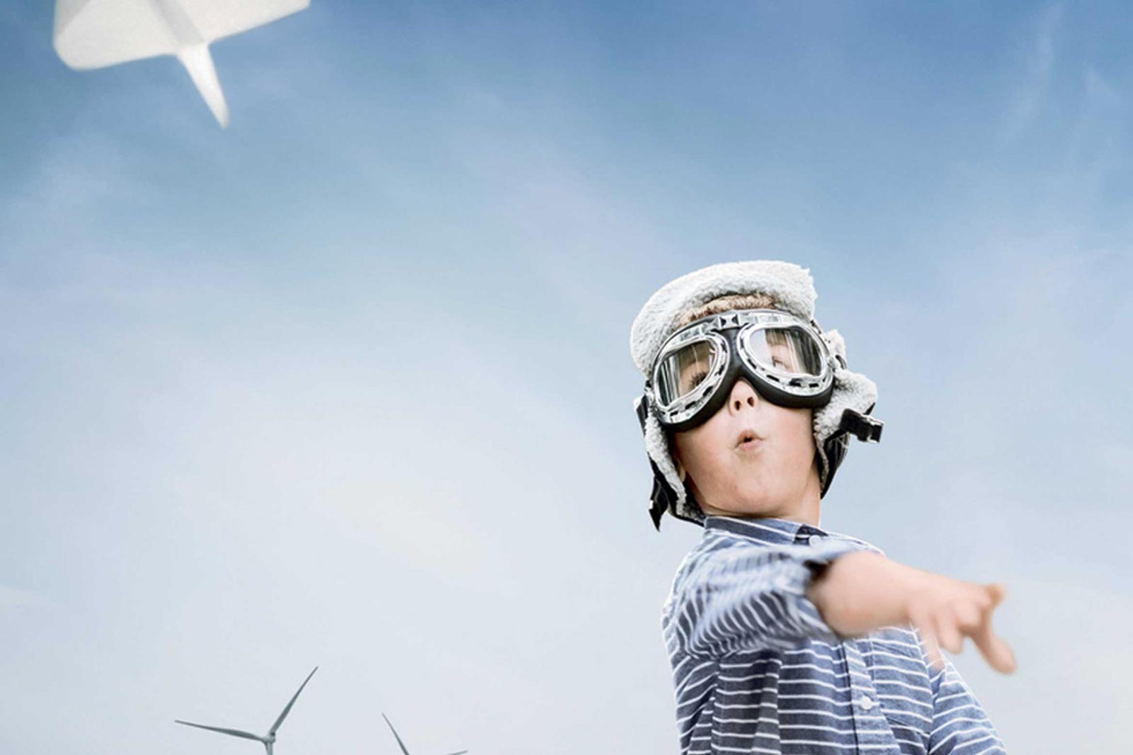 Ett barn i en randig skjorta pekar medan det bär en vit pilotmössa, med vindkraftverk i bakgrunden mot en blå himmel.