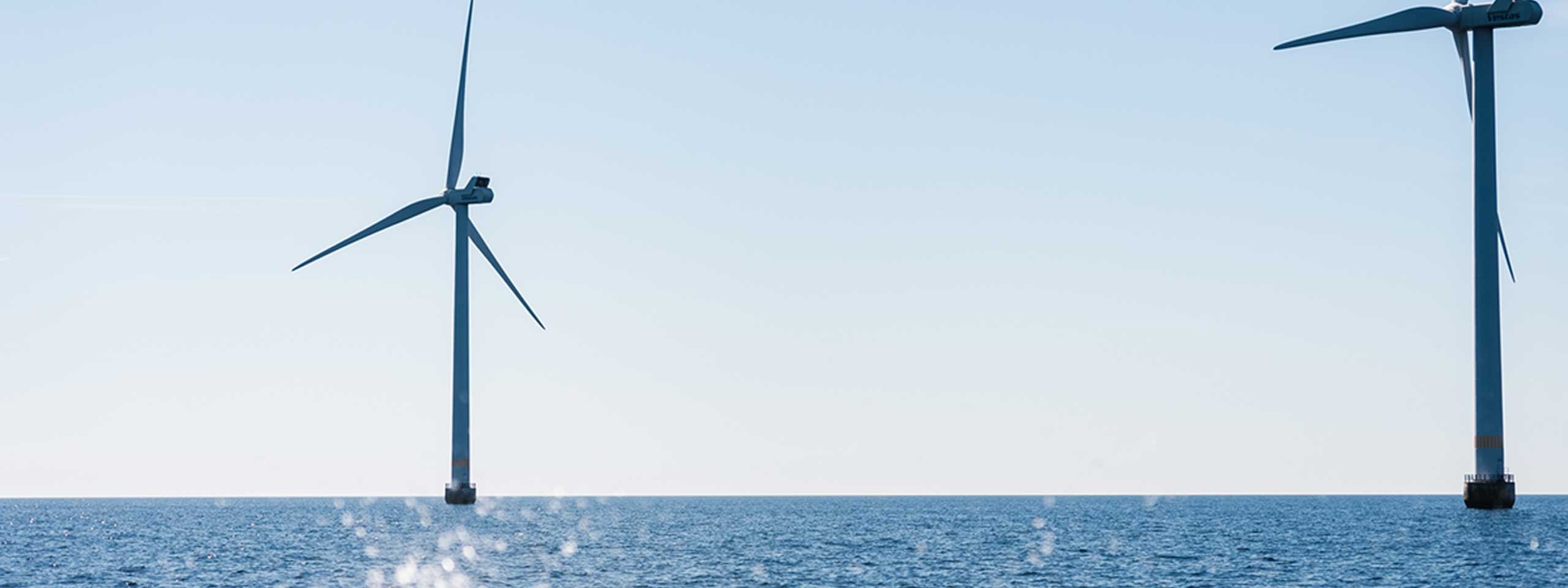 Two wind turbines stand in the sea under a light blue sky.