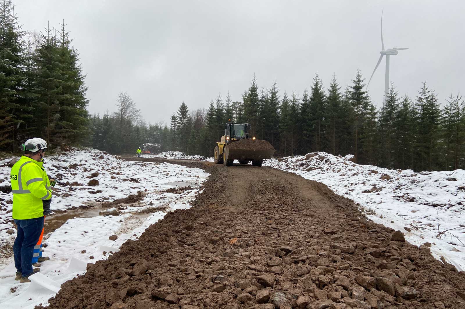 A construction site with a bulldozer moving earth on a snowy, muddy path in a forest with evergreen trees and a wind turbine.