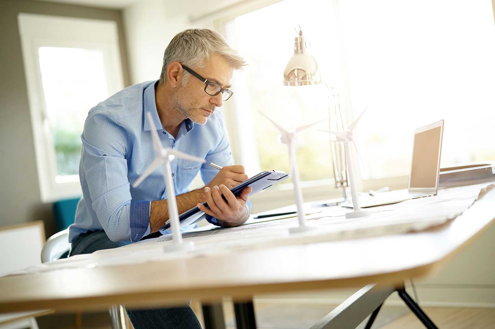 A man in a blue shirt is reviewing plans next to small wind turbines and a laptop in a bright, modern office.