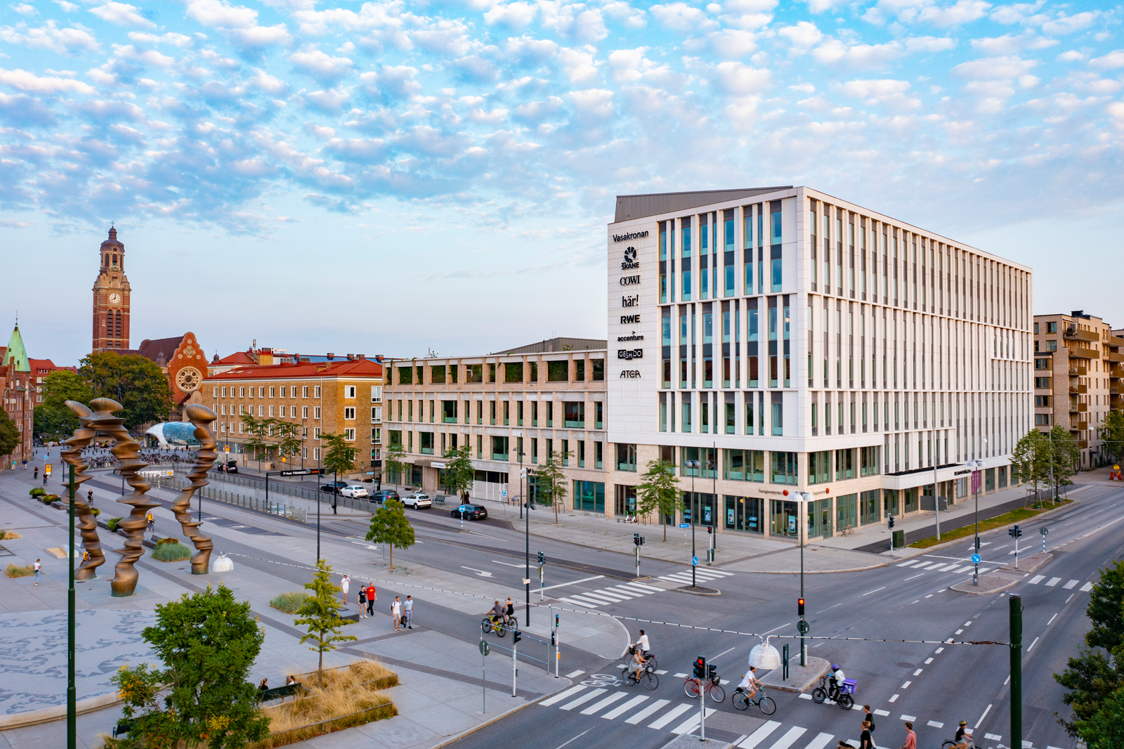 En stadsmiljö med moderna byggnader, cyklister och en stor skulptur i förgrunden. Himlen är klar med moln.