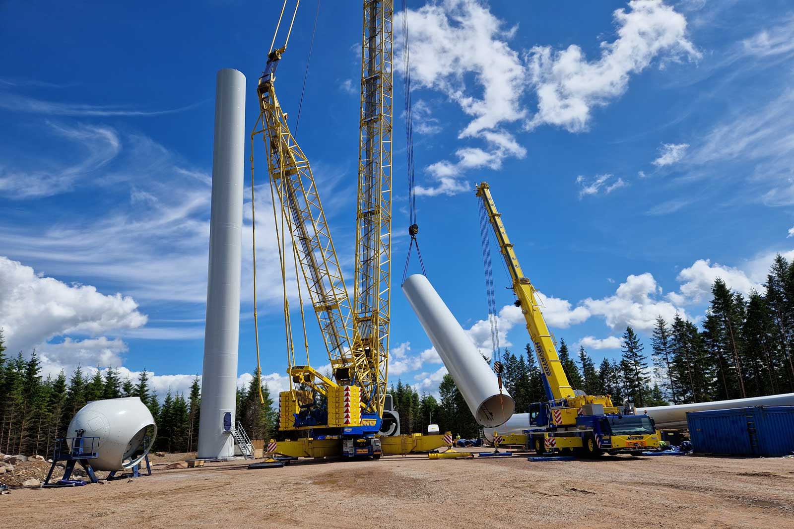En stor gul kran lyfter en vit turbinblad nära en grönskande skog under en klar himmel med moln.
