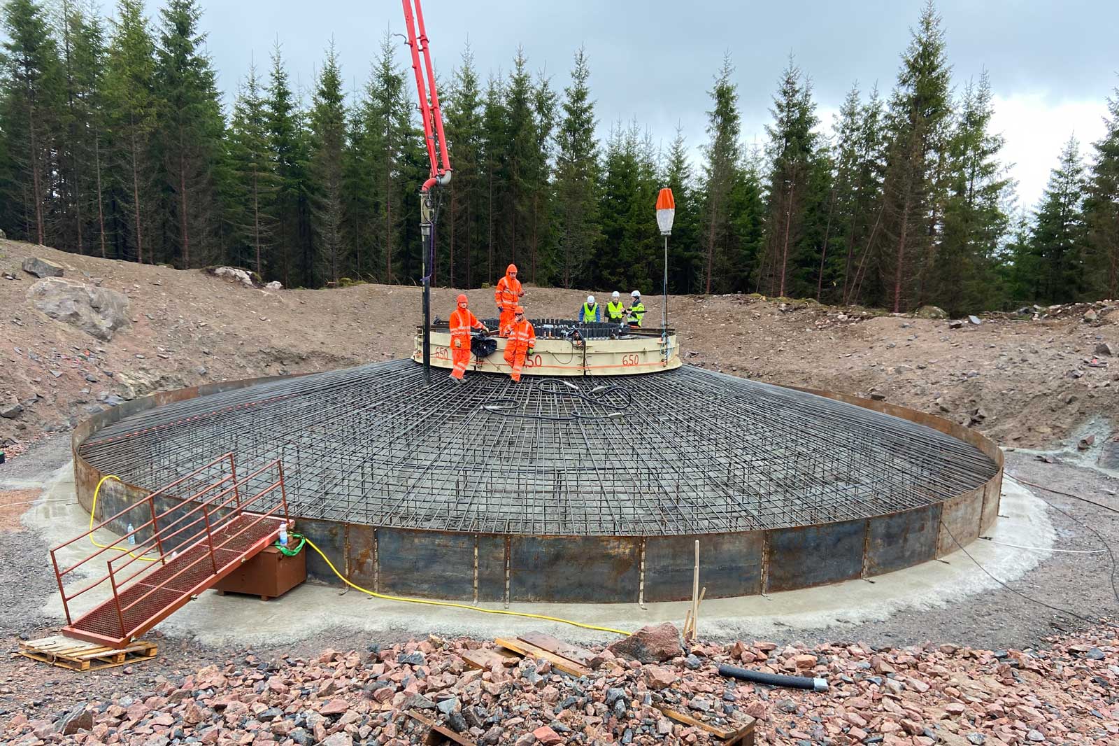 Arbetsplats för konstruktion av en stor betongstruktur i skogen. Arbetare i orangea kläder är aktiva i bygget.