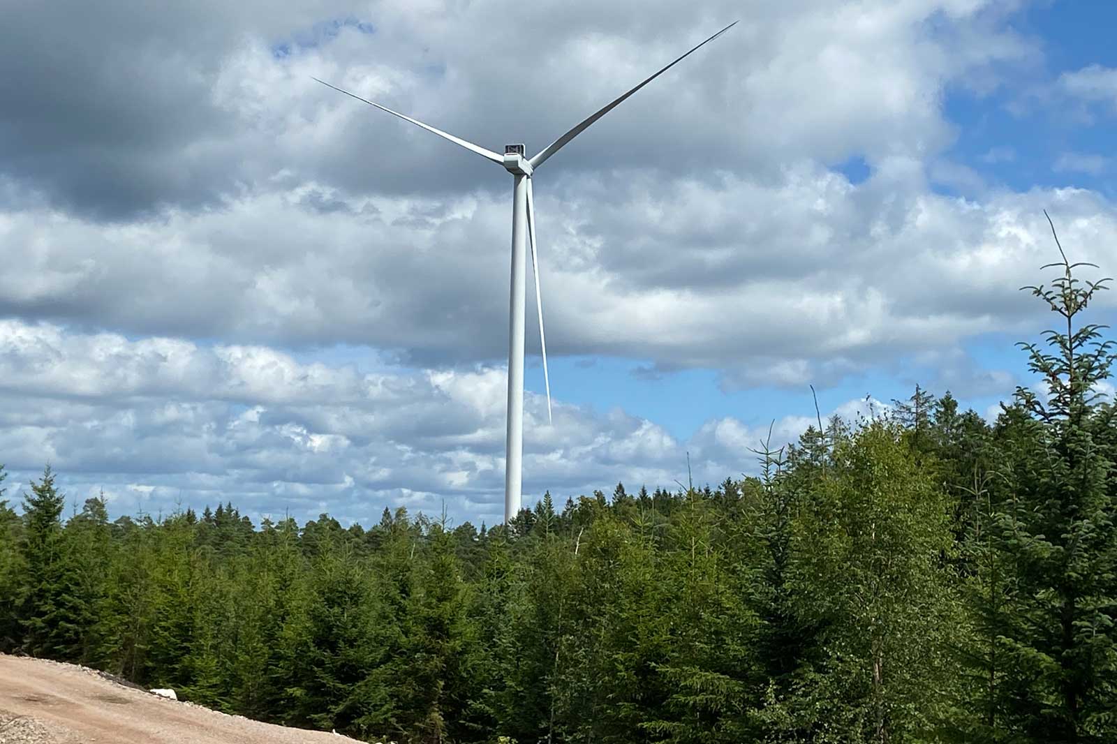 En hög vindkraftverk står mitt i en skog, under en molnig himmel med blåa partier.