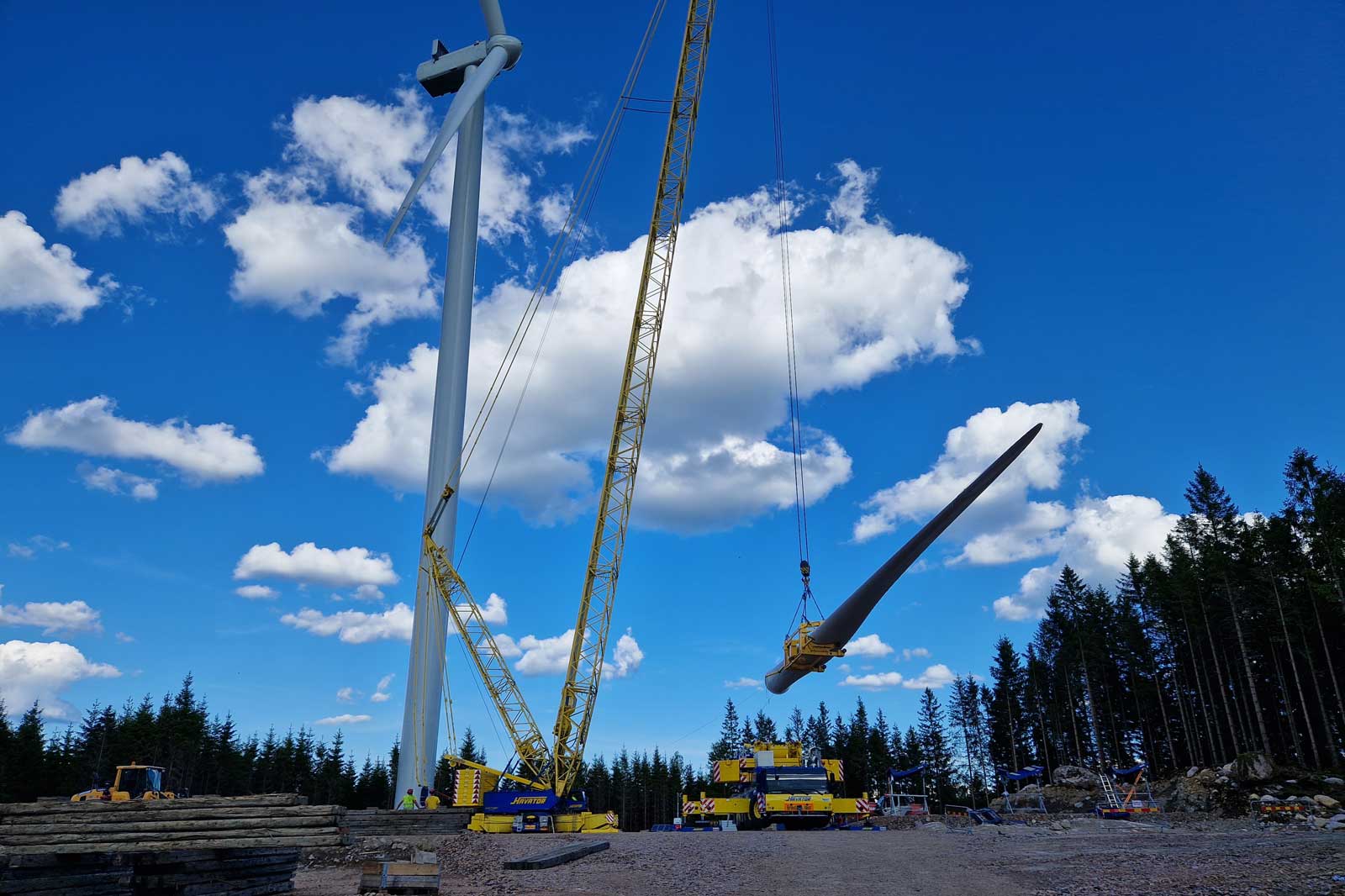 En kran lyfter en vindturbinblad under en blå himmel med vita moln. Skog och byggmaskiner syns i bakgrunden.