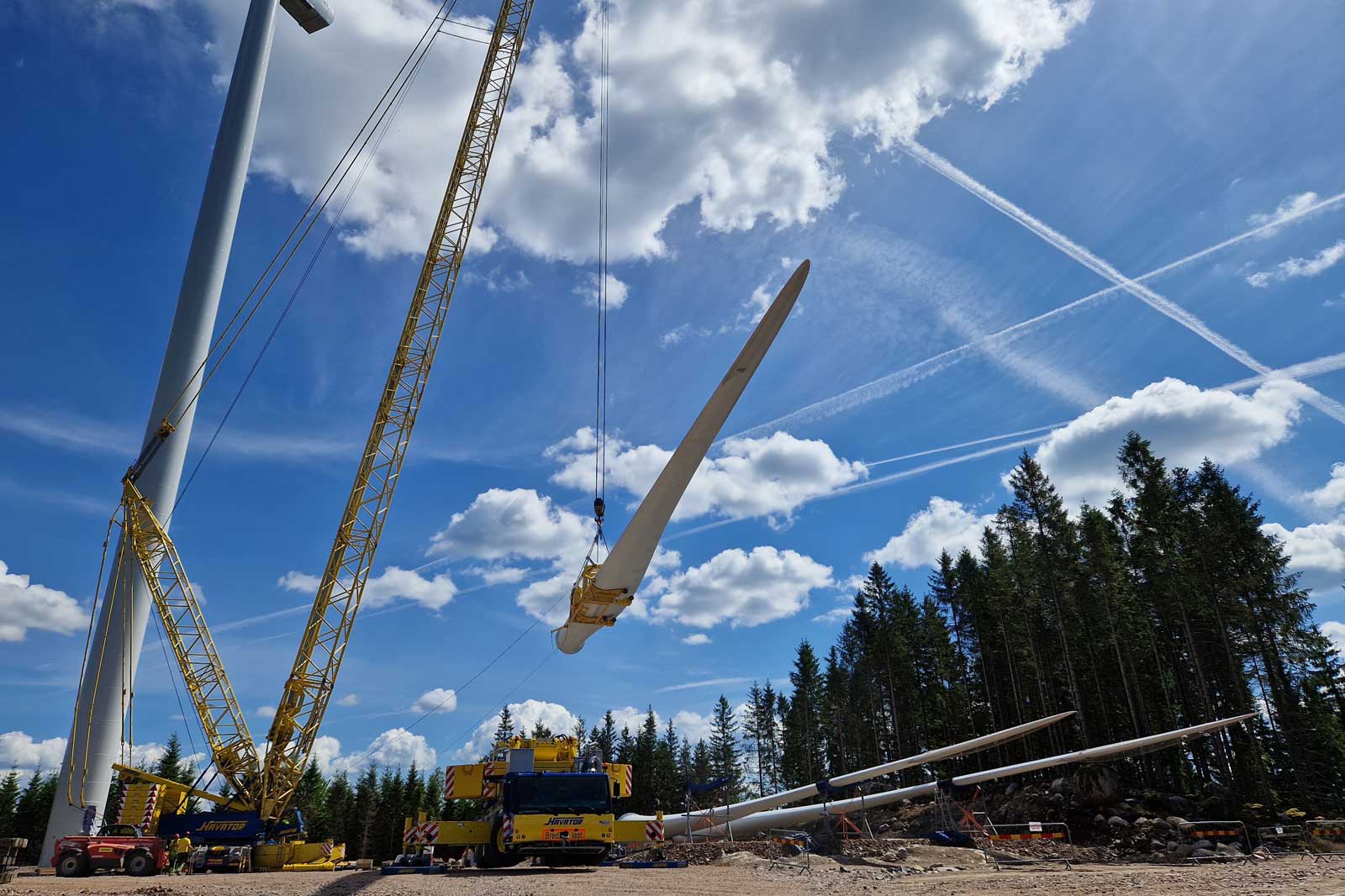 En stor kran lyfter en vindkraftsblad mot en blå himmel med fluffiga moln och en skog i bakgrunden.