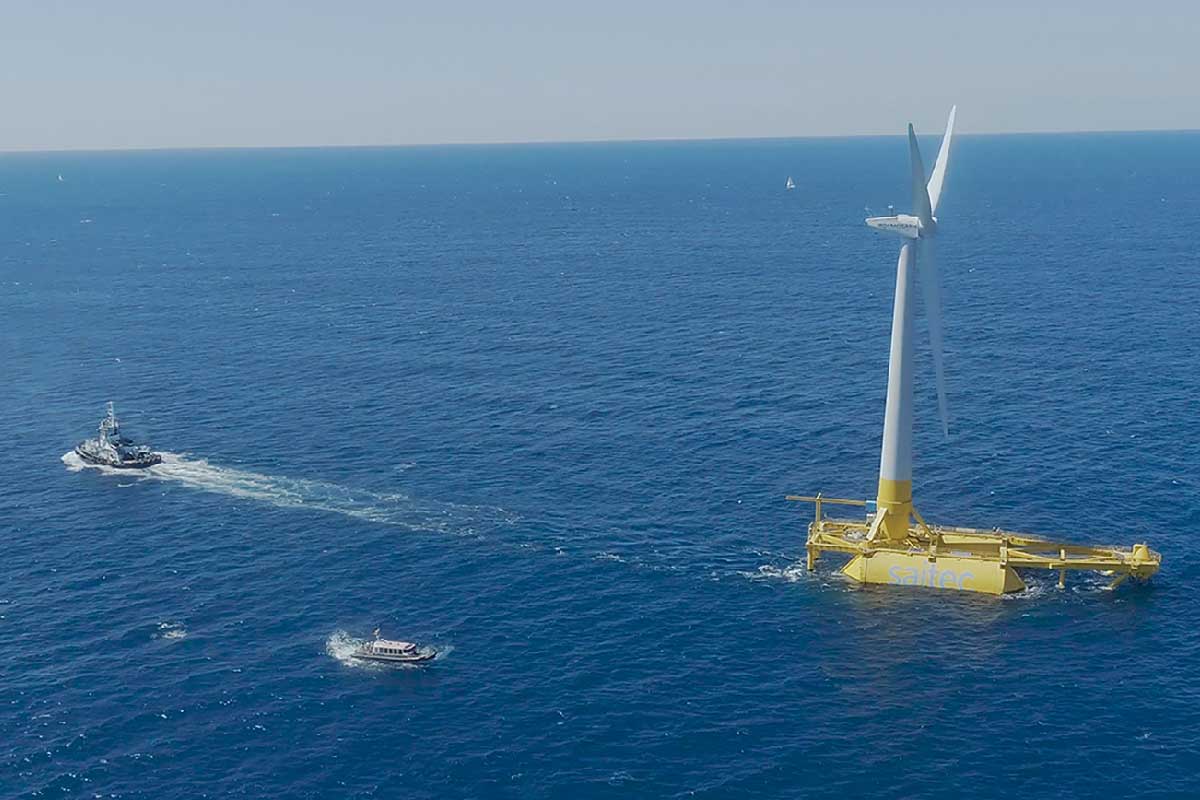 A view of an offshore wind turbine anchored in deep blue waters, with two boats nearby.