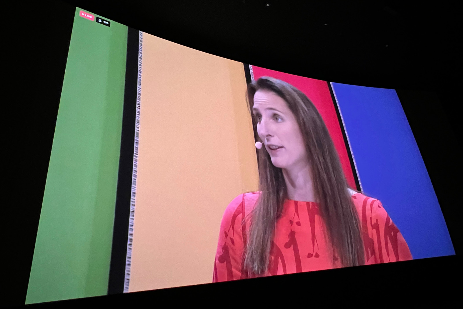 En kvinna i en livlig röd klänning står på scen med en färgglad bakgrund av gröna, gula och blå paneler.