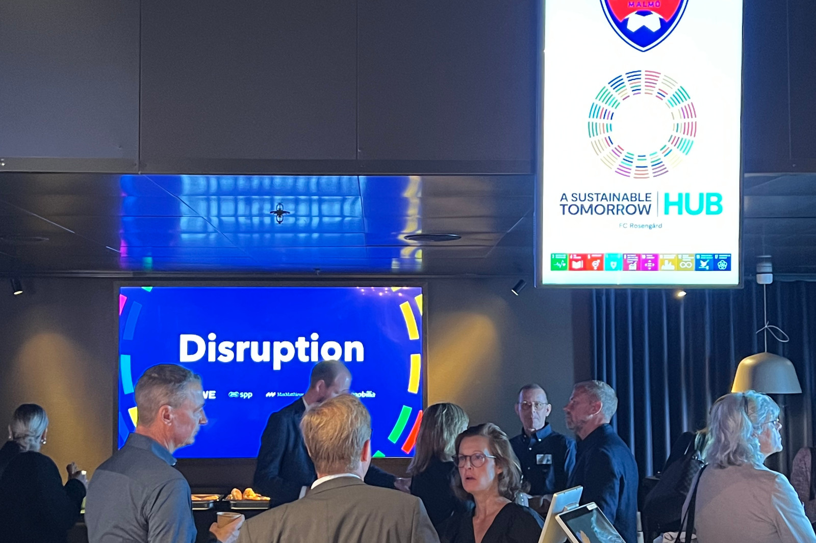 En grupp människor interagerar i en modern miljö, med en skärm som visar ordet 'Disruption' och en hållbarhets-hub skylt.