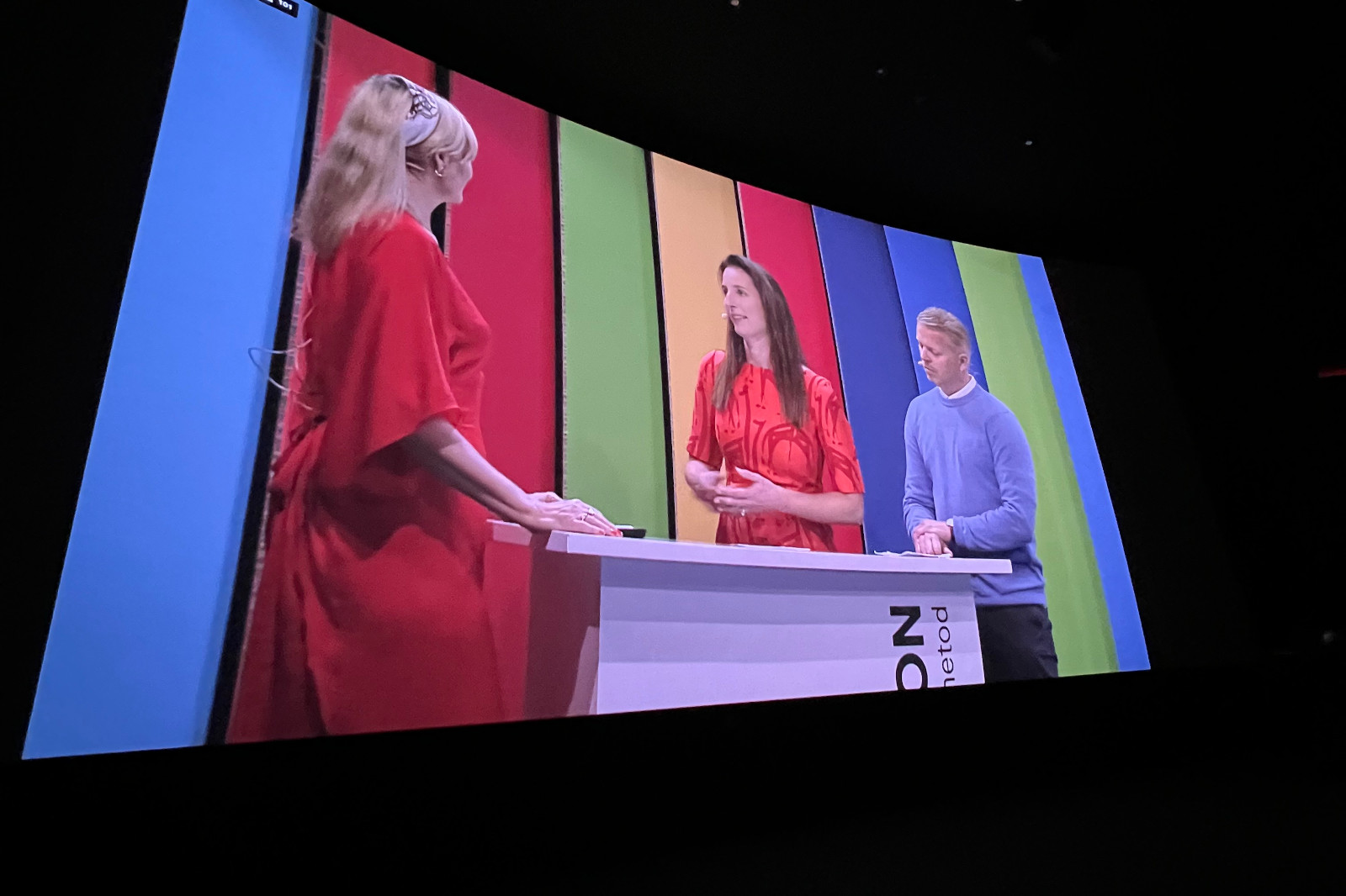 En livlig tv-studioscen med tre presentatörer vid ett bord framför färgglada bakgrundspaneler.