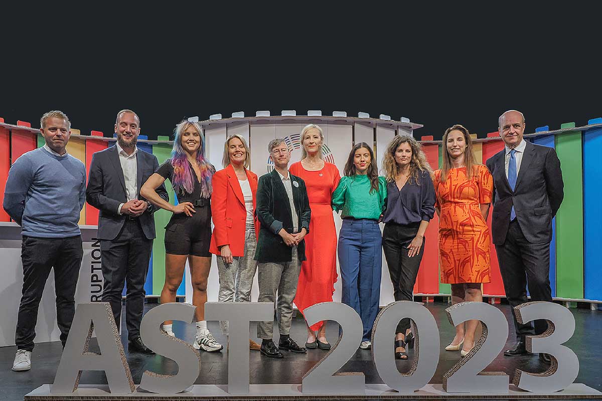 Grupp av nio personer som står på en scen med en färgglad bakgrund och texten 'AST 2023' i förgrunden.