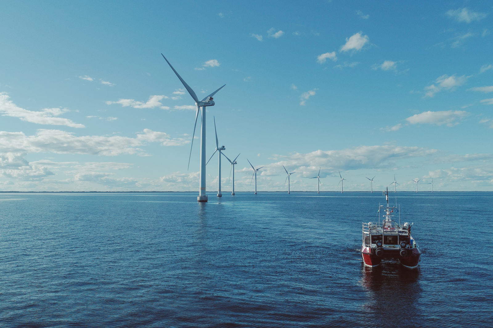 En båt seglar i lugna vatten nära vindkraftverk under en klarblå himmel med spridda moln.