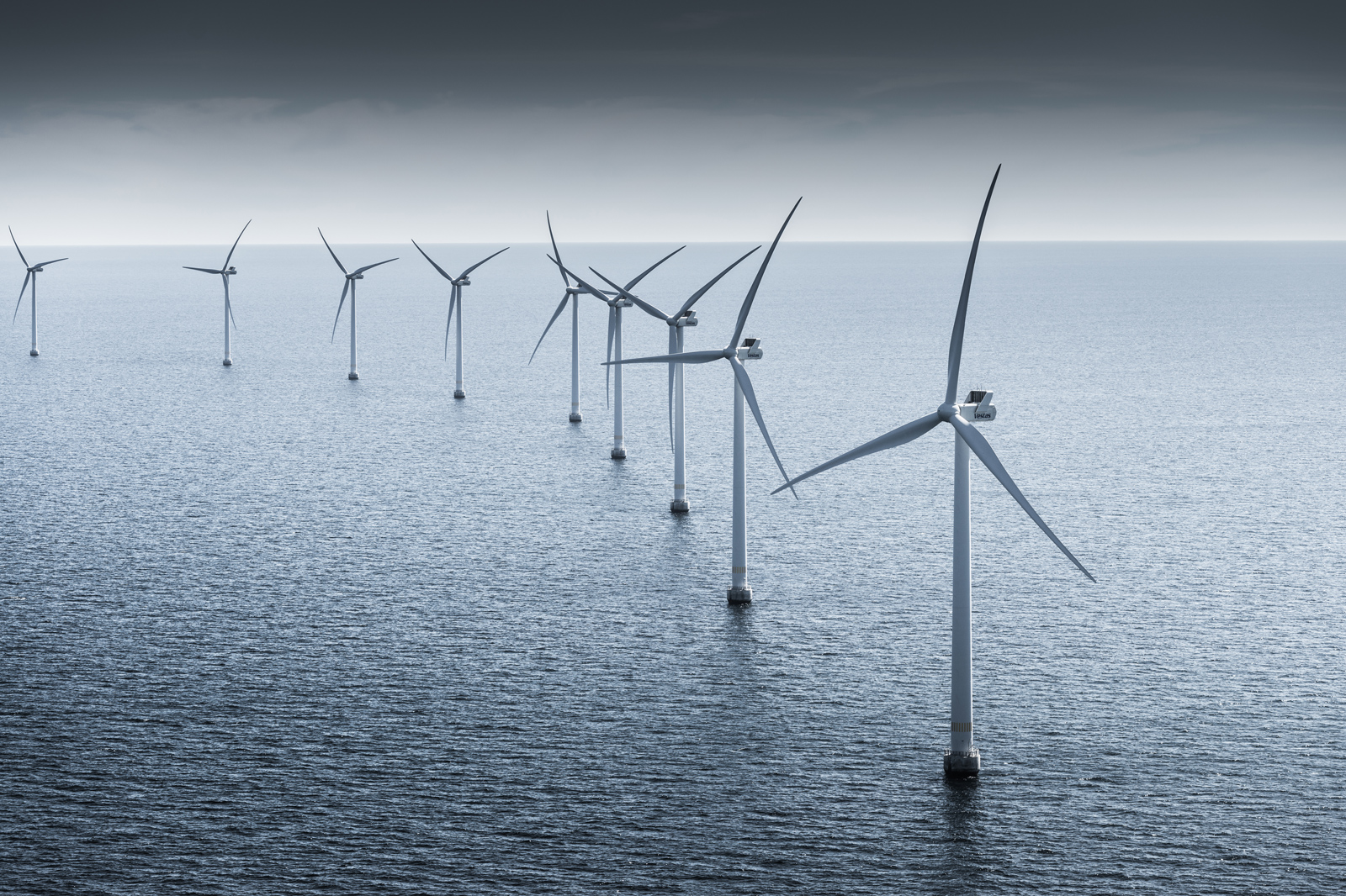 En rad av vindturbiner som står i havet, med en molnig himmel ovanför och lugnt vatten nedanför.