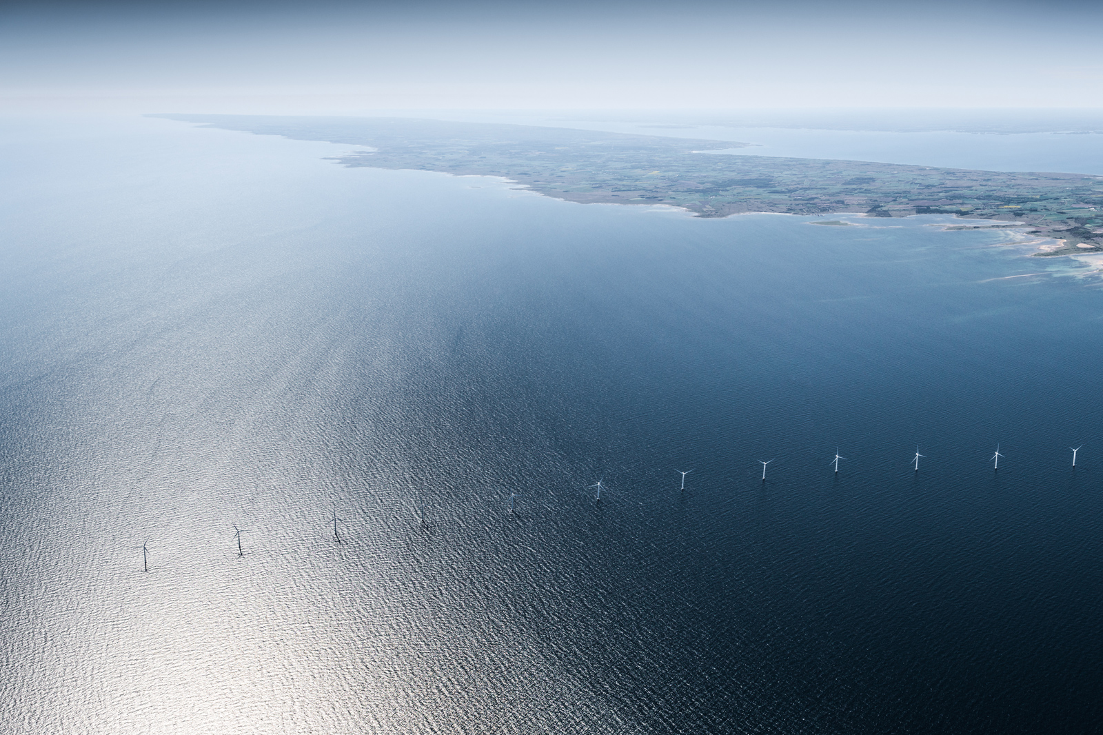 Fågelperspektiv över vindkraftverk i havet, med en kustlinje synlig i fjärran.