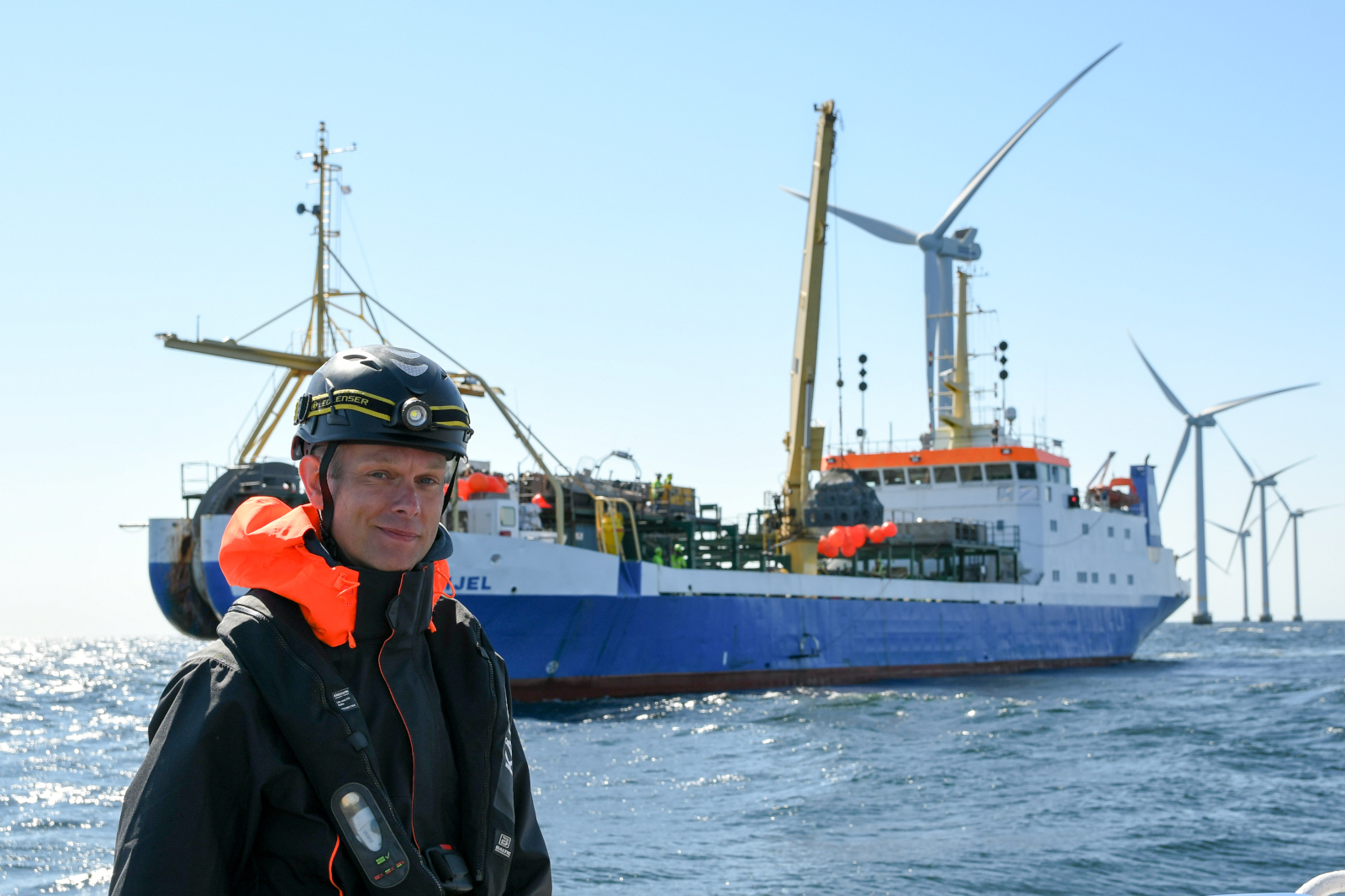 En person i en säkerhetsdräkt framför ett fartyg nära vindkraftverk på havet.