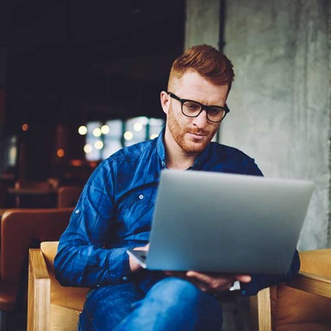 En person som sitter i ett café, använder en bärbar dator i en avslappnad outfit, mjukt ljus skapar en avkopplande atmosfär.