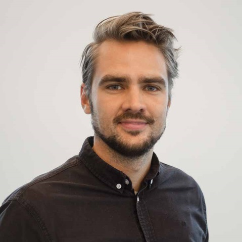 En person som bär en mörk knappad skjorta poserar mot en enkel vit bakgrund.