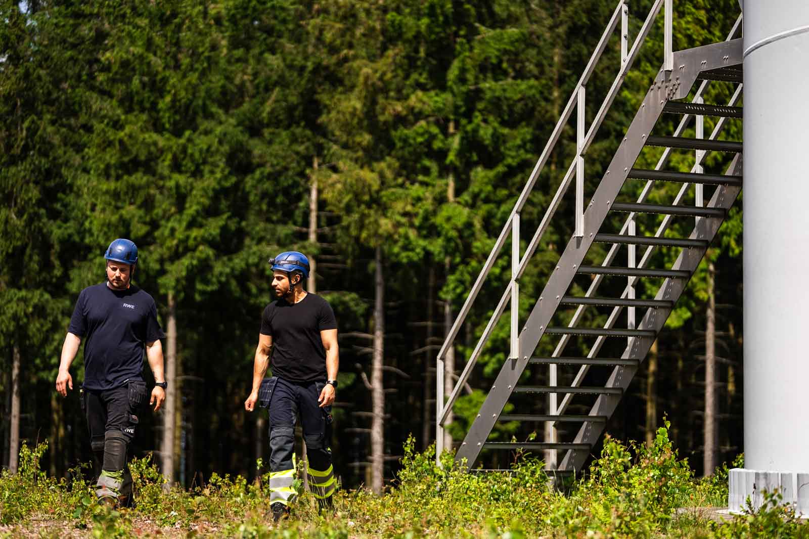 Två arbetare i hjälmar går mot en vindturbin, omgivna av träd och gräs.