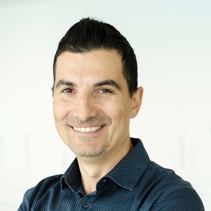 En man i en mörk mönstrad skjorta står mot en ljus bakgrund, med armarna i kors.