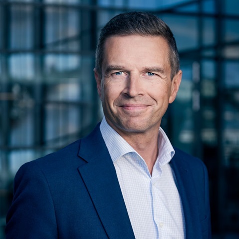 En man i en blå kostym och vit skjorta står utomhus mot en glasbyggnad i bakgrunden. Porträtt av Axel Alm.