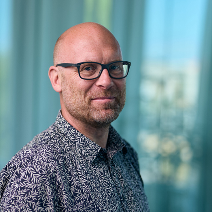 En man med glasögon som bär en blommig skjorta, står framför genomskinliga gardiner i en ljus inomhusmiljö.