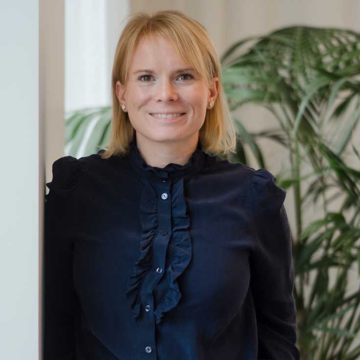 En kvinna i en mörk, volangblus står bredvid en pelare i ett rum med gröna växter i bakgrunden.