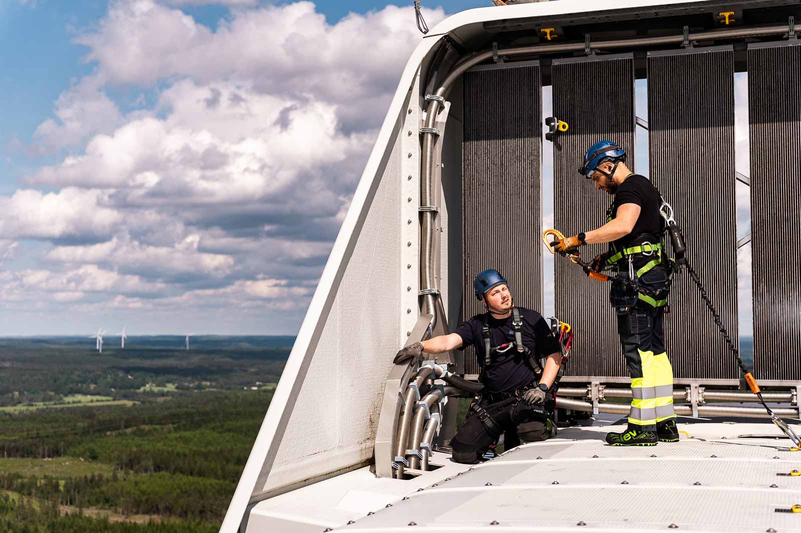 Två arbetare i säkerhetsutrustning utför underhåll på en vindkraftverk. De omges av en naturskön landskap.
