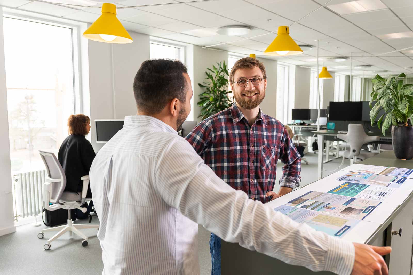 Två män samtalar i ett modernt kontorsutrymme med ljusa, gula lampor och gröna växter, medan dokument ligger utspridda på ett bord.