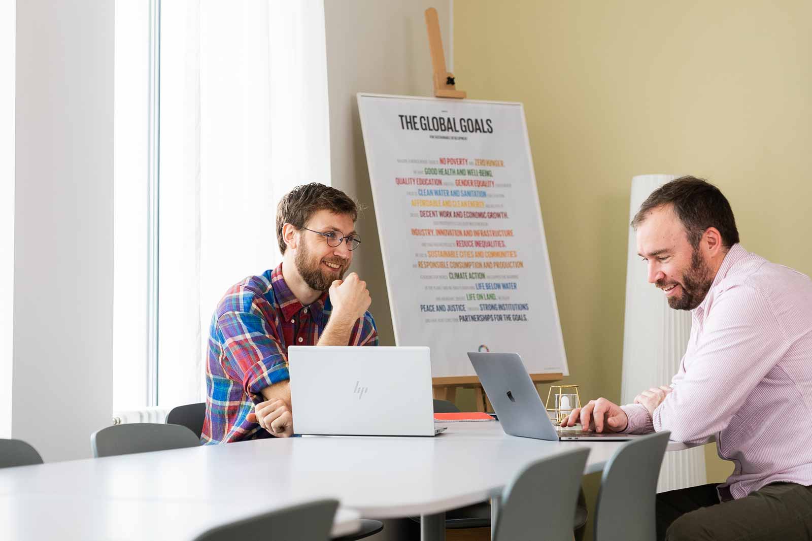 Två män diskuterar vid ett bord med bärbara datorer, en affisch om globala mål i bakgrunden.
