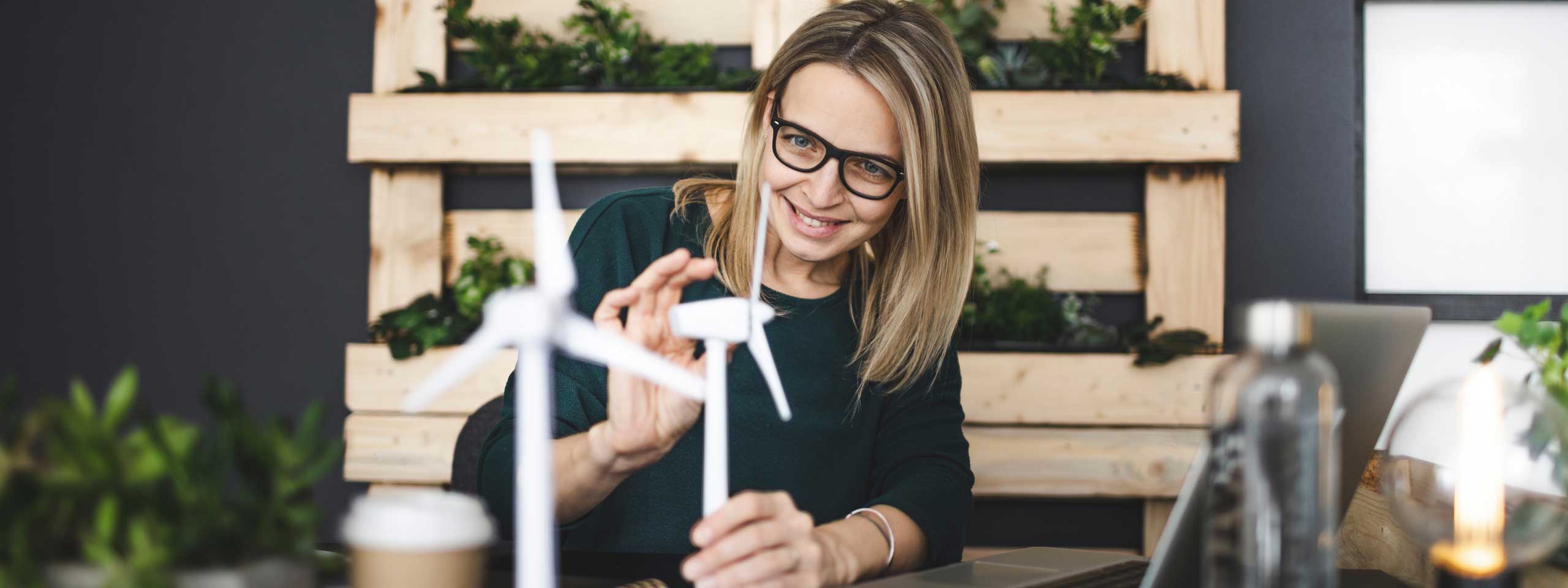 A woman is working on a small wind turbine model at a desk surrounded by plants, with a laptop and a coffee cup nearby.