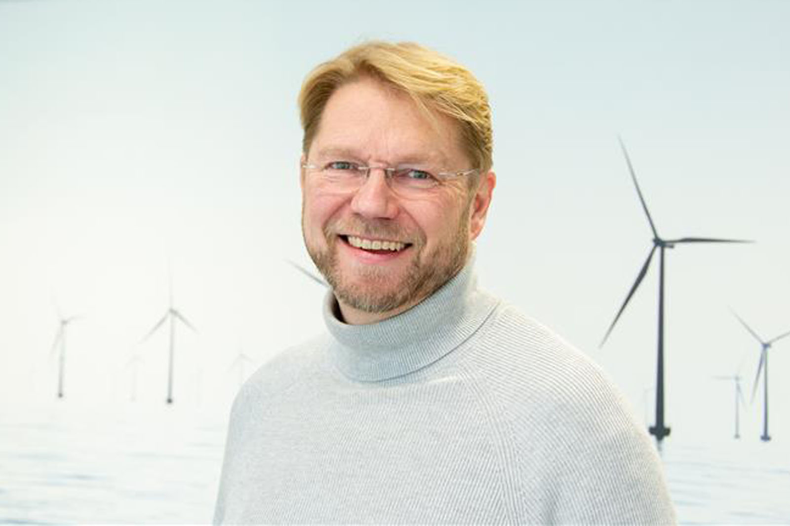 En person som bär en grå polotröja står mot en bakgrund av vindkraftverk i ett stillsamt landskap. Porträtt av Fredrik Uggendahl.