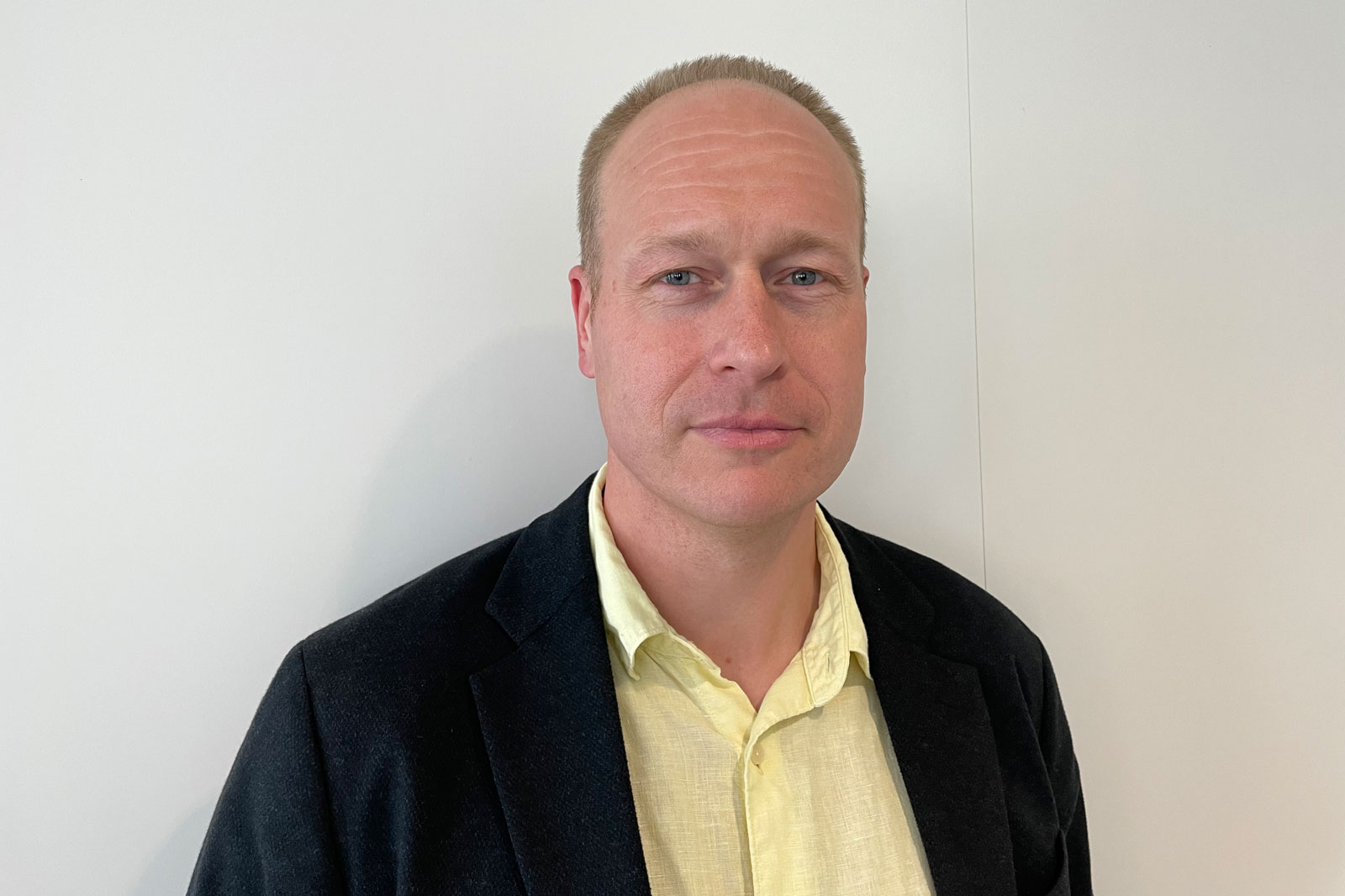 En person i en svart kavaj med en gul skjorta, poserande framför en enkel vit bakgrund.