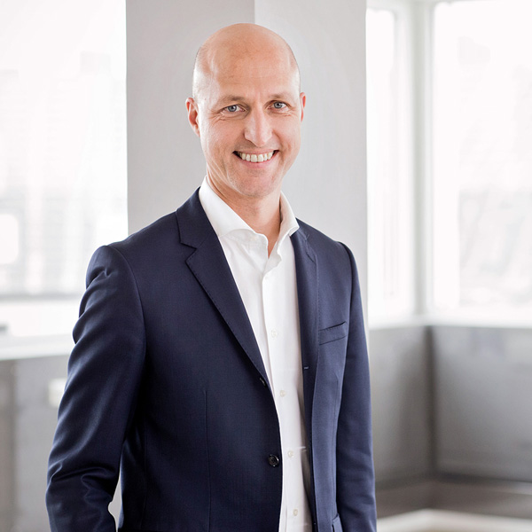 A man in a dark blue suit stands in a modern office with large windows, portrait of Sven Utermöhlen.