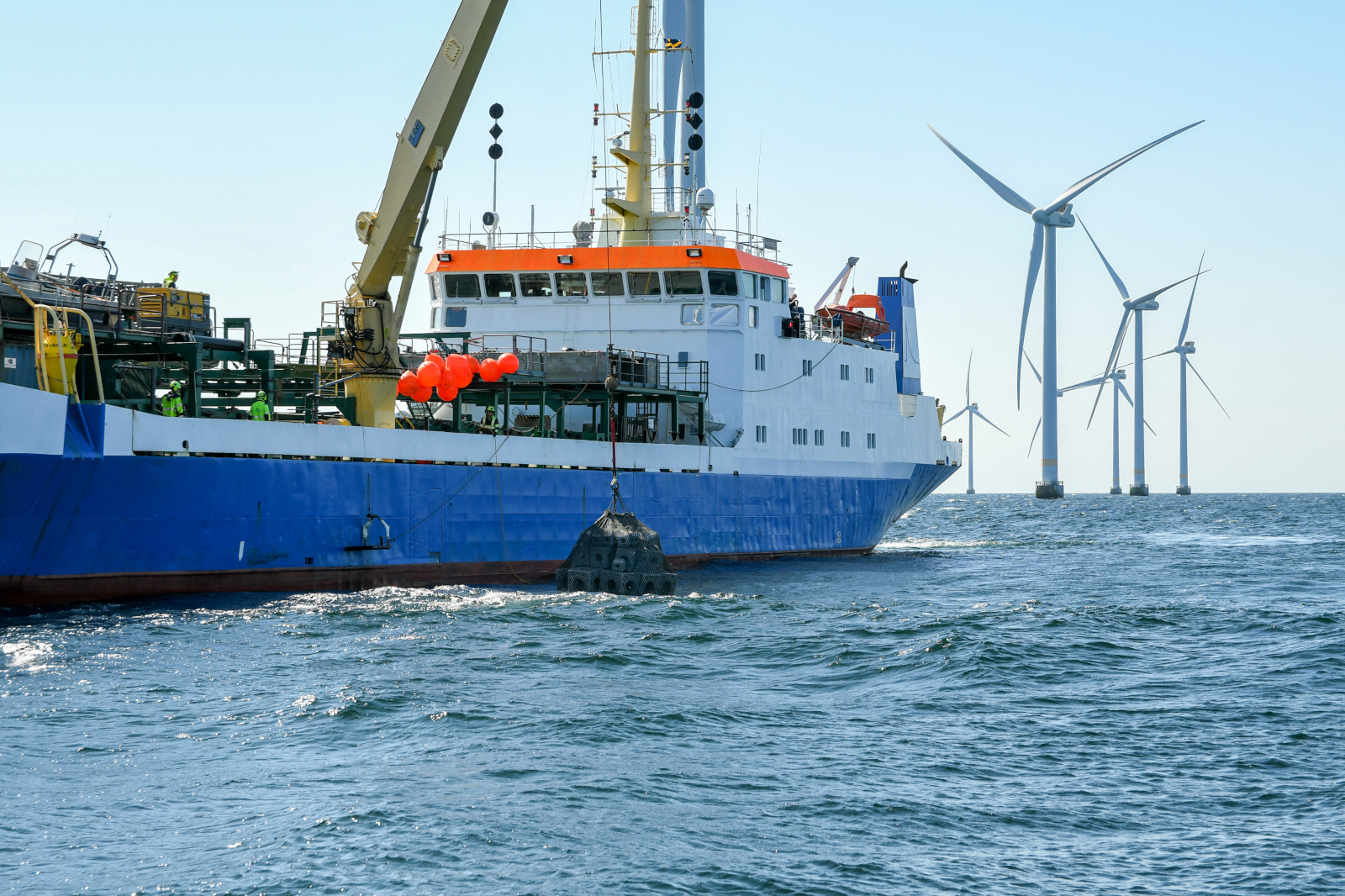 A blue and white ship with balloons berths near wind turbines in the sea, showcasing marine operations and renewable energy.