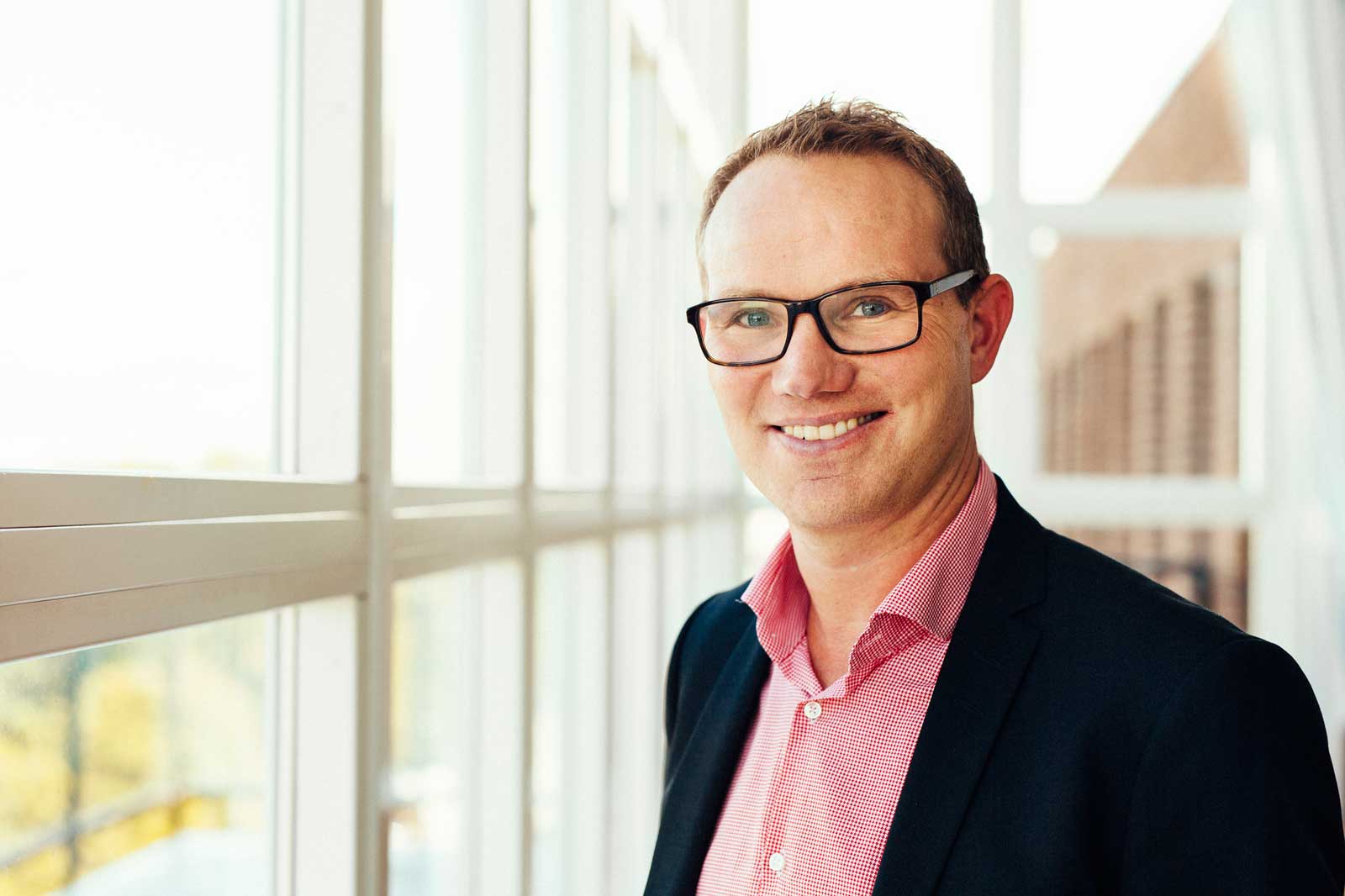 En professionellt klädd person står bredvid stora fönster, med naturligt ljus som belyser scenen.