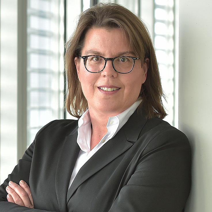 A person in a black suit stands with crossed arms in front of modern windows with lots of light. Portrait of Regina Wolter.
